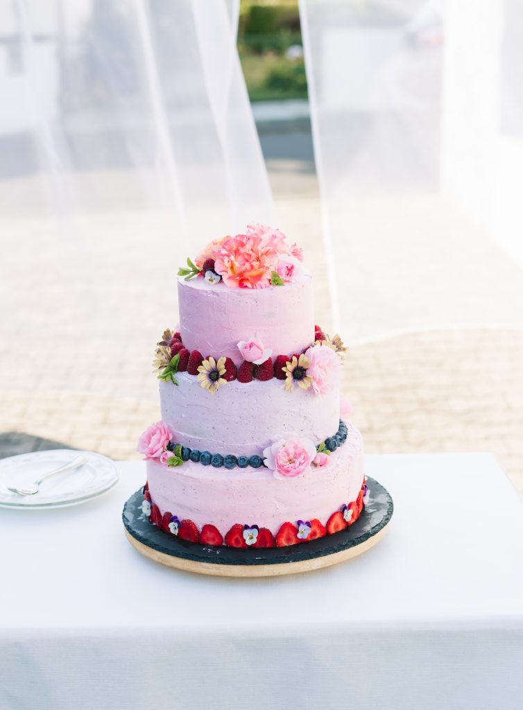 Eine dreistöckige vegane Hochzeitstorte steht auf einem Tisch auf einer Schifferplatte. Viele essbare Blüten sind auf der Sahnecreme angebracht. Erdbeeren, Himbeeren und Heidelbeeren umranden jeweils einen Stock der Torte und zeigen so an welcher Geschmack sich darin verbirgt.