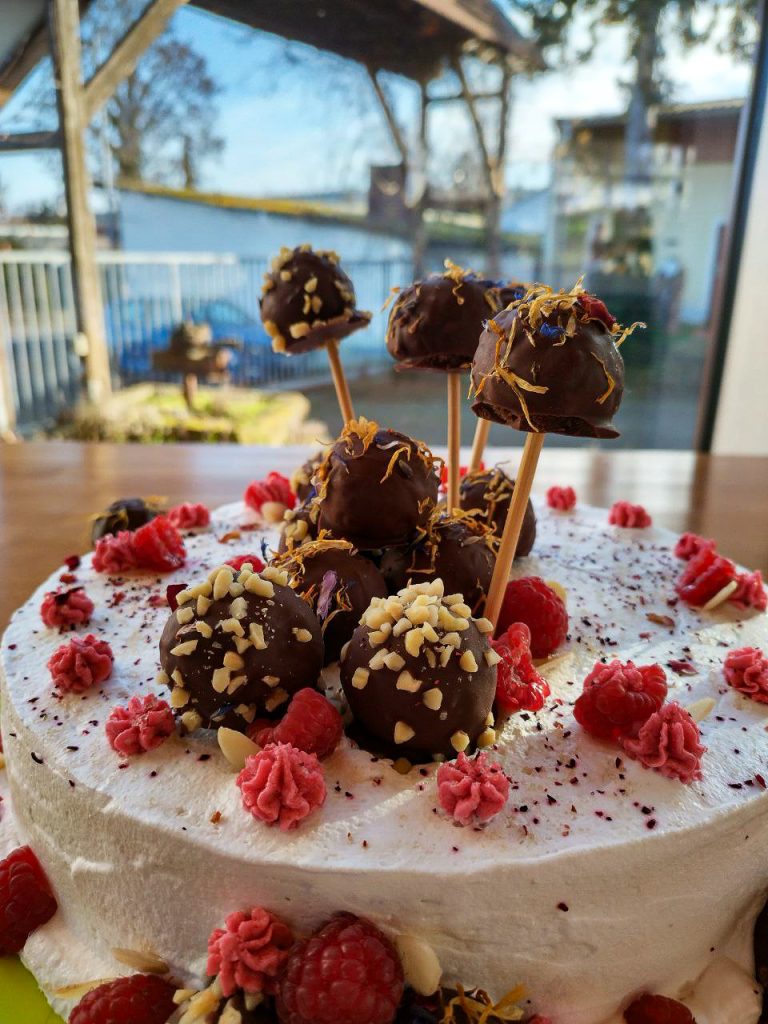 Eine vegane Himbeertorte mit Cakepops dekoriert steht auf einem Tisch. Die Torte wirkt farbenfroh und üppig. Zusätzlich ist sie mit Mandeln, Himbeeren, und rosa Buttercreme verziert.