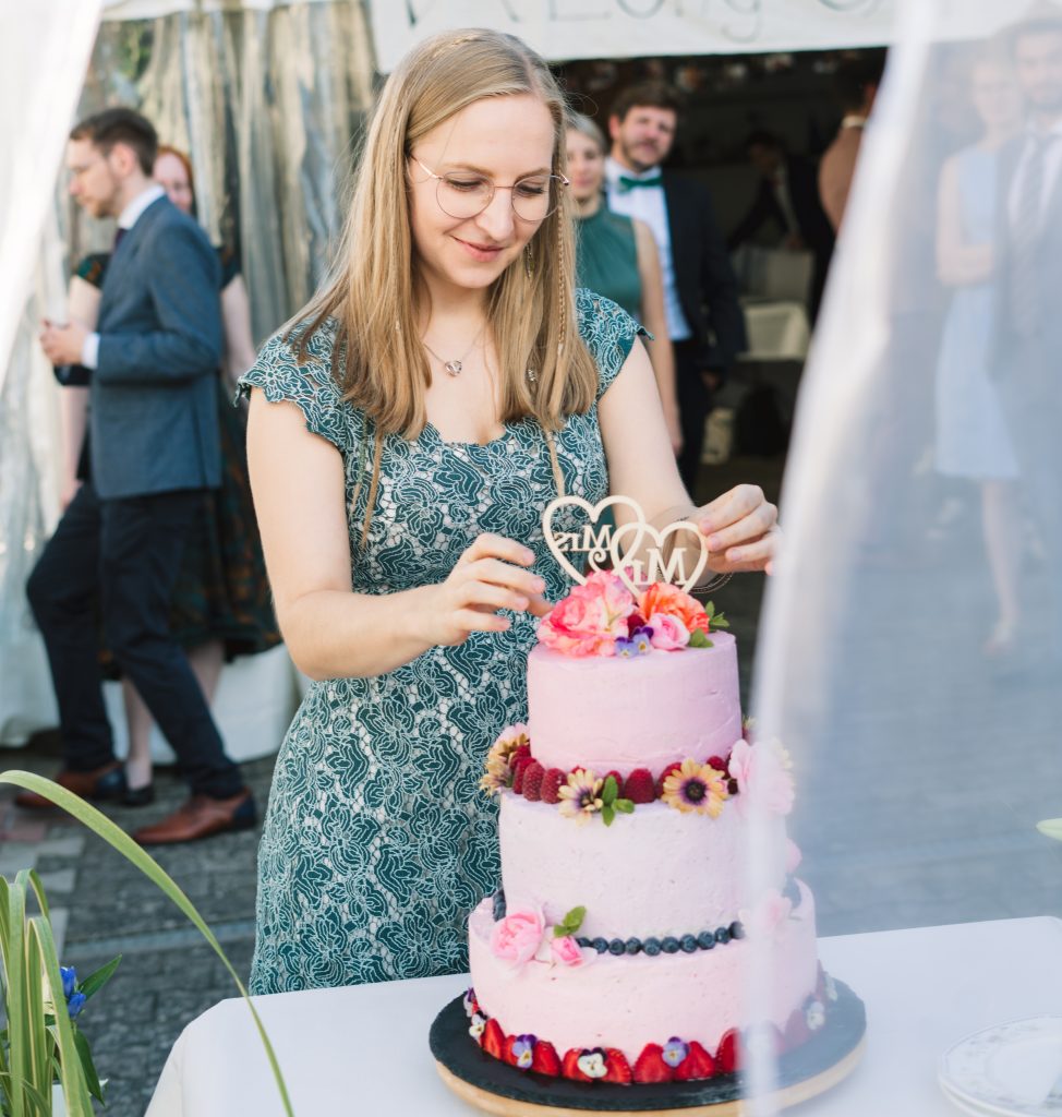 Irina, die Inhaberin des Wombats Cafés steht vor einer für Freunde gebackenen veganen Hochzeitstorte. Sie setz die letzten Dekoelemente auf die, mit Blumen und frischen Früchten verzierte Torte und lächelt.
